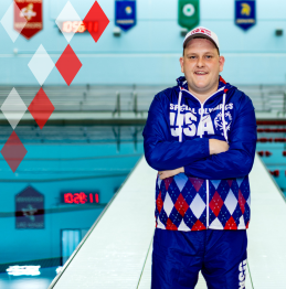 Matthew standing in front of an indoor swimming pool and wearing the official gear for the United States Special Olympics team.