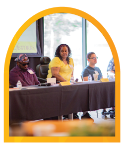 Picture of Kiesha, wearing a yellow t-shirt and sitting at a long table, with panelists on either side of her at our recent Disability Inclusion Breakfast event. Picture is surrounded by a yellow and orange border.
