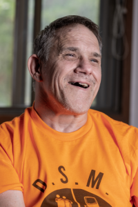 Photo of Michael wearing his orange Disabled Speech Matters t-shirt and smiling in his apartment.