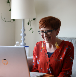A photo of Jessica, a woman with red-brown hair and wearing glasses, sitting on a couch and smiling as she looks at her computer.