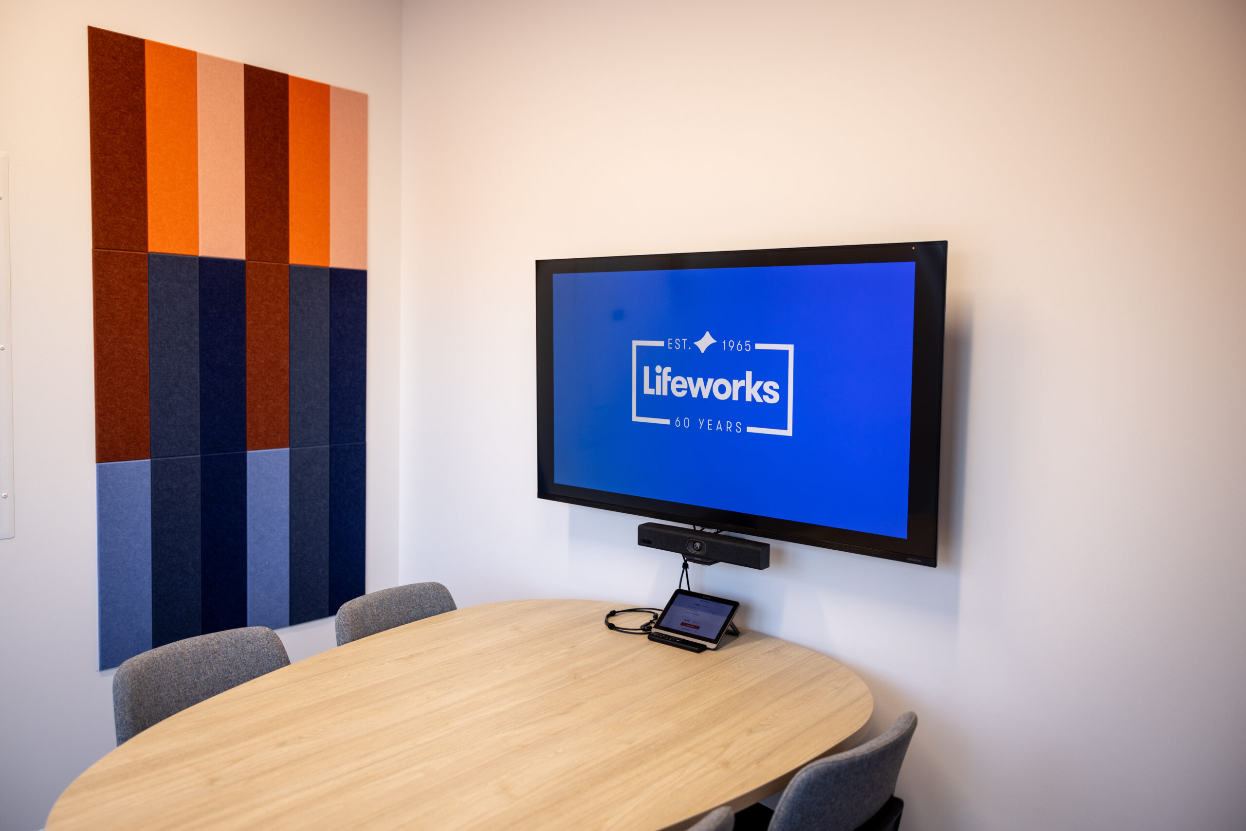 Focus room at the Lifeworks Center for Disability Inclusion in New Hope. Shows a table with four chairs around it, with a television screen mounted on the wall above the table. On the television screen is the Lifeworks logo. On the wall adjacent to the screen are red, orange, pink, and blue felt tiles for decoration.